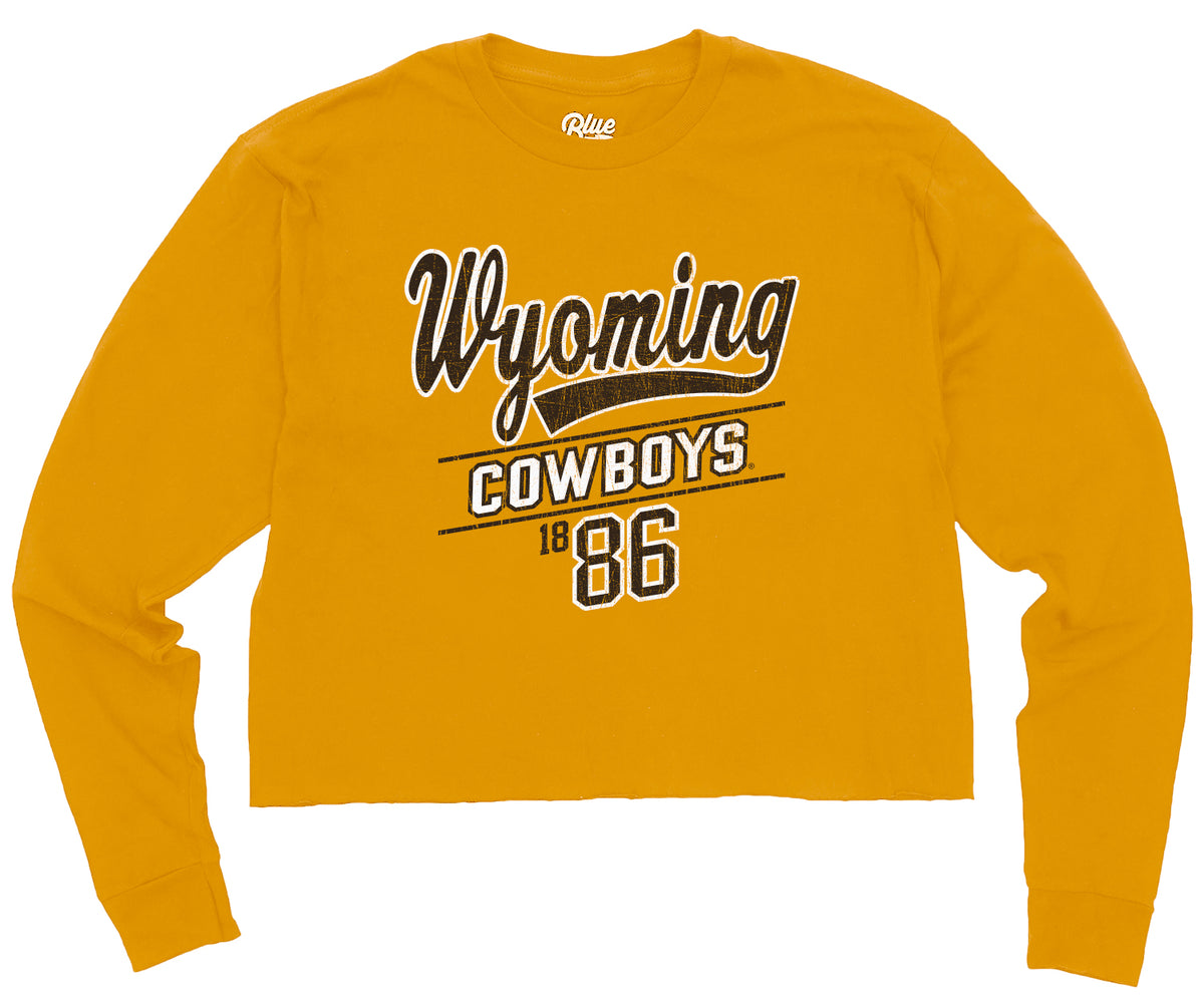 gold womens crop long sleeve with design centered. design is brown wyoming script with white outline, with cowboys in white below and brown 1886 below that