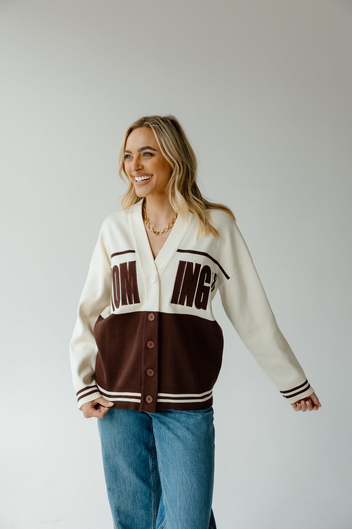A woman modeling a varsity-style cardigan with a cream upper section, dark brown lower panel, and dark brown lettering on the chest, paired with blue jeans against a light gray studio background.