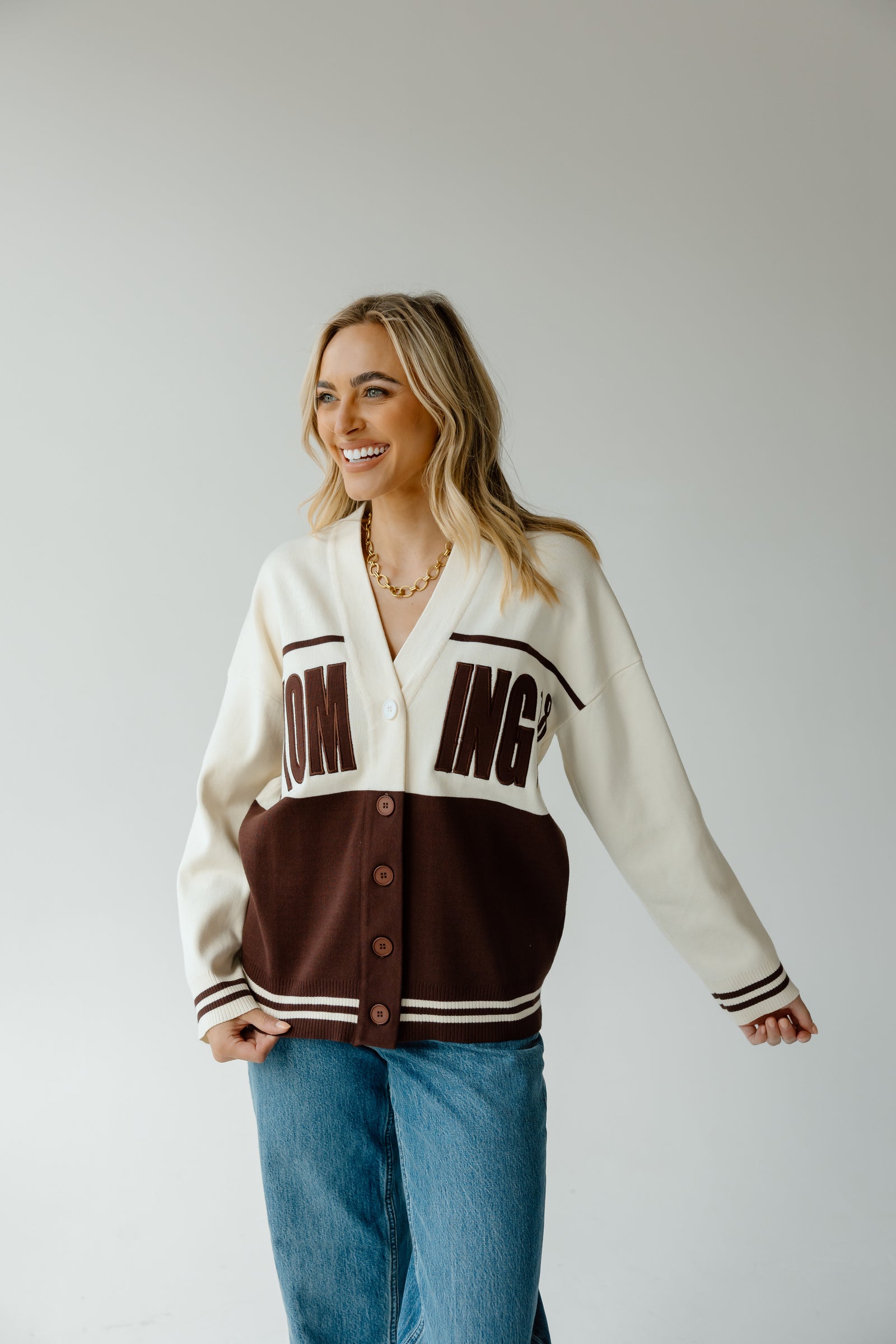 A woman modeling a varsity-style cardigan with a cream upper section, dark brown lower panel, and dark brown lettering on the chest, paired with blue jeans against a light gray studio background.