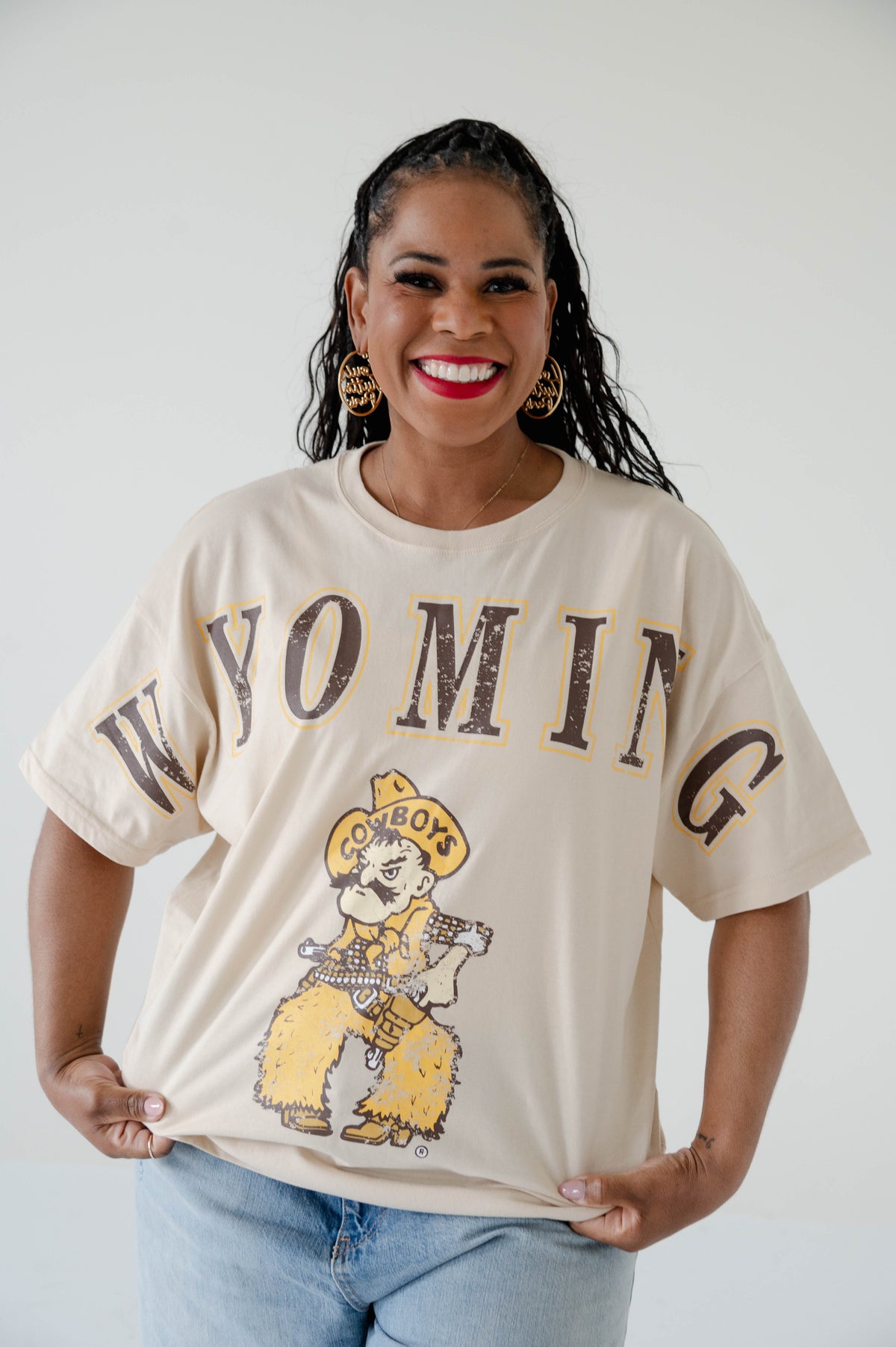 Woman posing wearing a oatmeal shirt with wyoming and pistol pete in gold and brown all over the front.