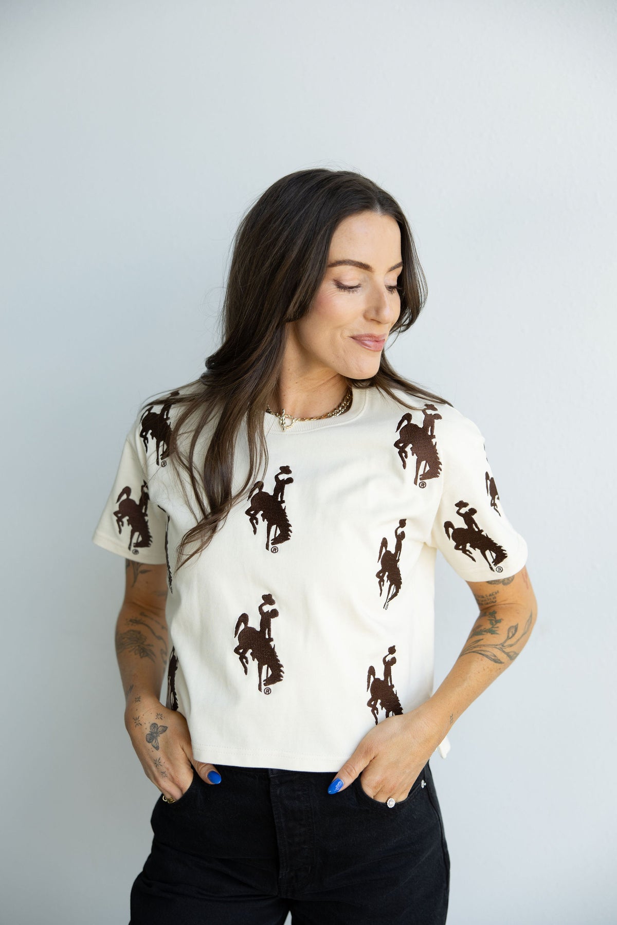A woman wearing a cream-colored short-sleeve t-shirt with a dark brown bucking horse print scattered all over, paired with black jeans, standing against a light gray studio background.