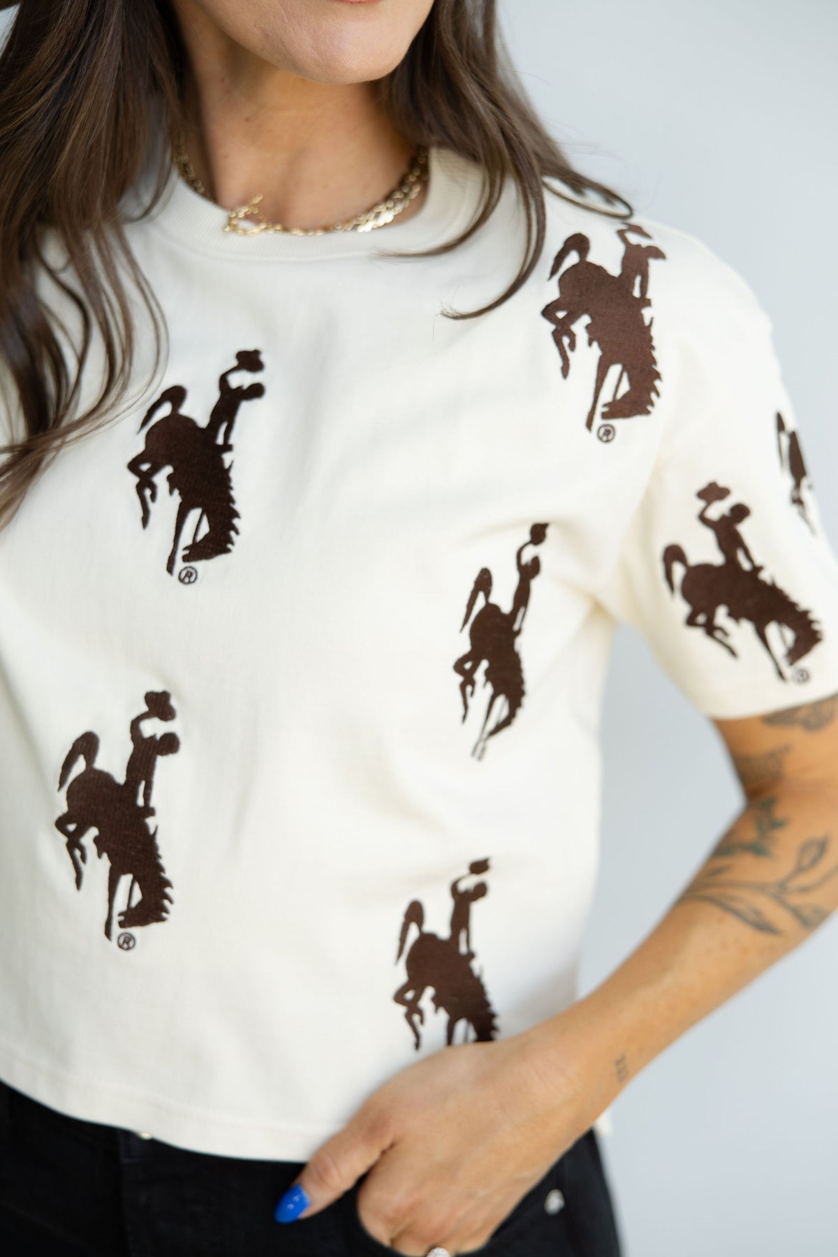 A close-up of a person wearing a cream-colored short-sleeve t-shirt with an allover dark brown bucking horse. The person is also wearing a gold chain necklace and black pants, with blue nail polish on their hand.
