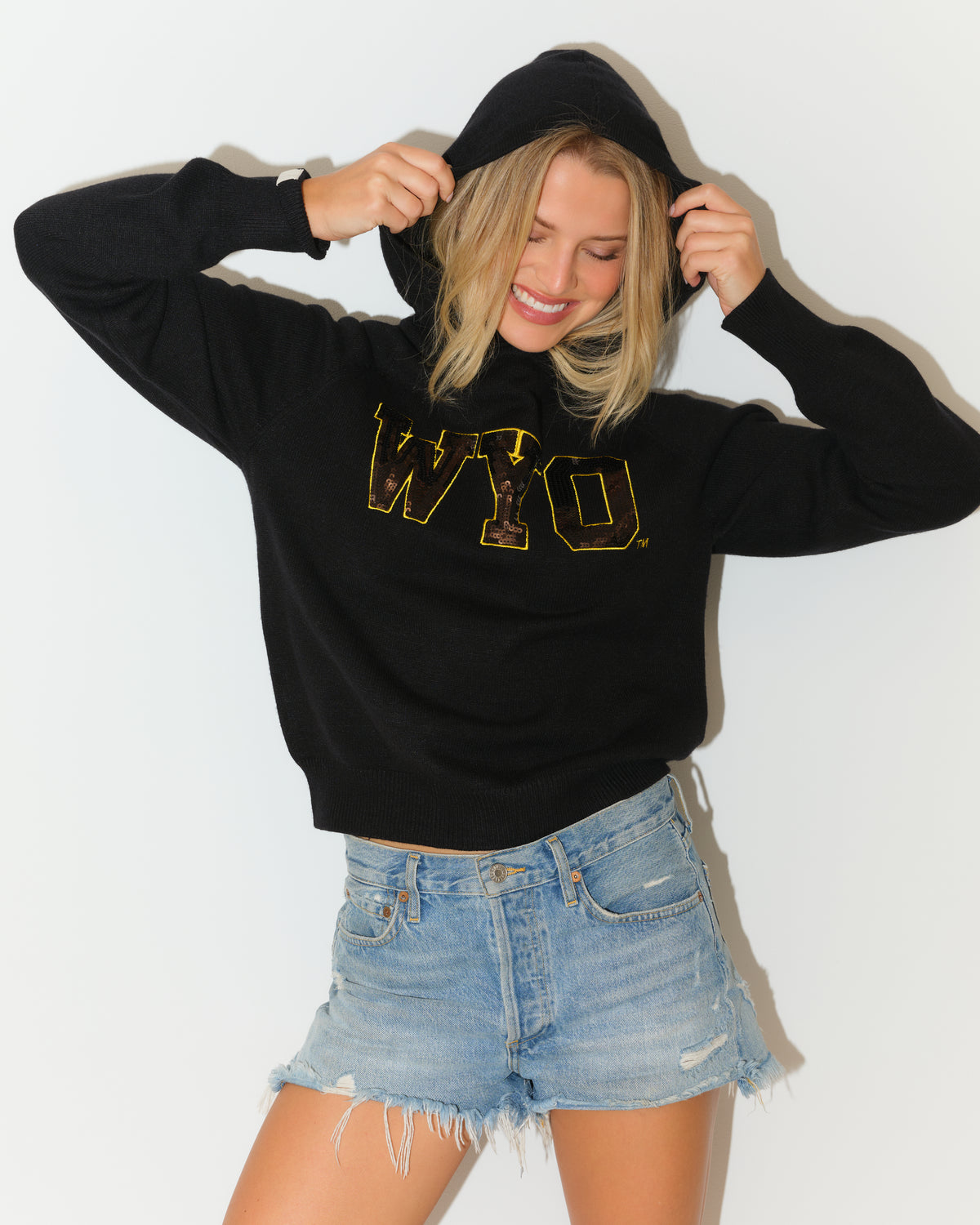 Woman modeling black hoodie pullover with wyo in brown sequin with gold outline wearing blue jean shorts. Pictured on a white background.