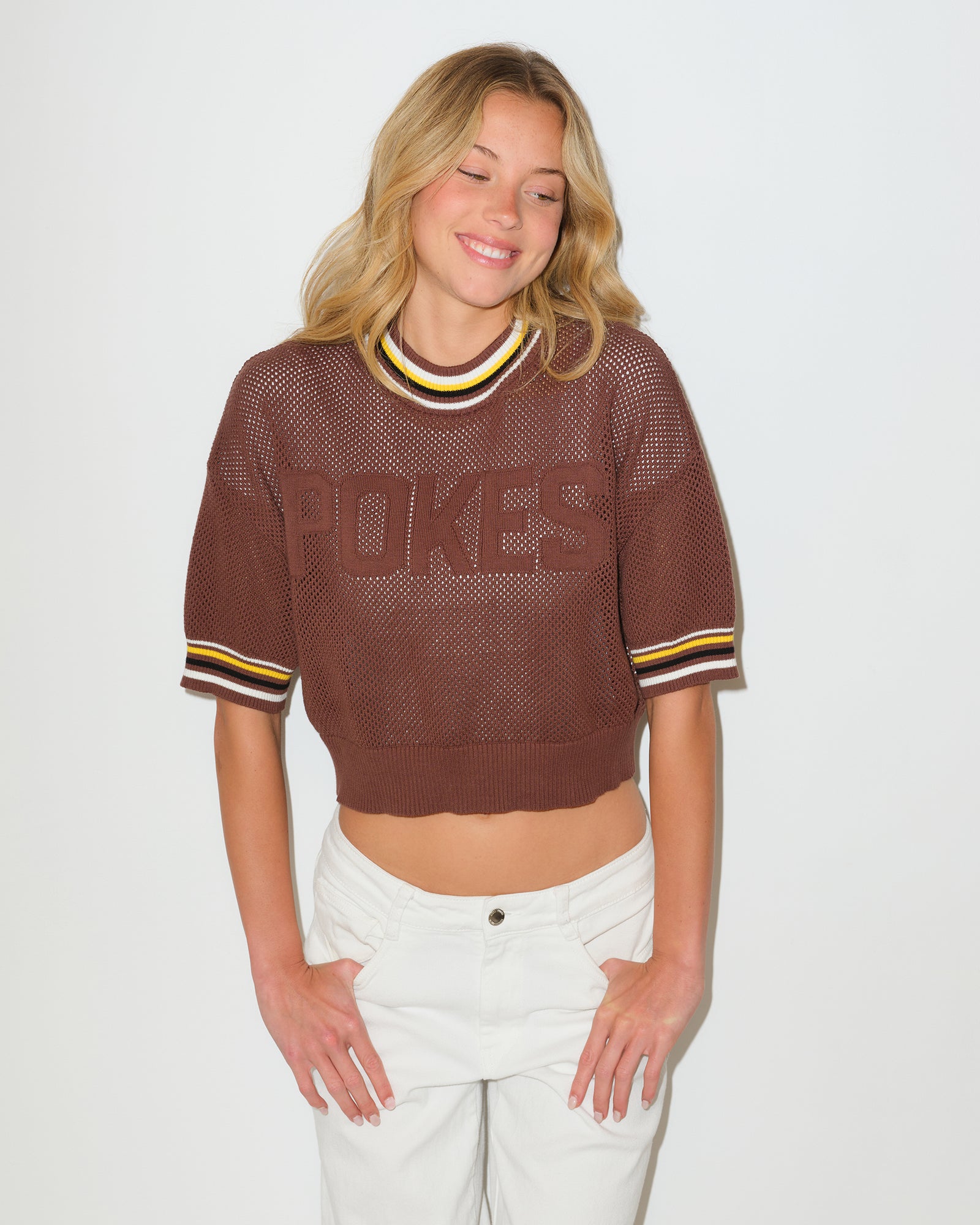 Woman modeling a short sleeve brown sweater that has gold white and black pinstripes on collar and sleeves. Is see through and has pokes lettering in brown on front chest. Pictured on a white background.