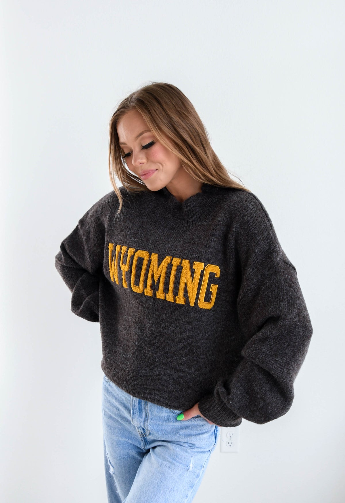 A woman wearing a dark brown fuzzy knit sweatshirt with 'WYOMING' in bold gold varsity lettering across the chest, paired with light blue jeans, set against a plain white studio background.