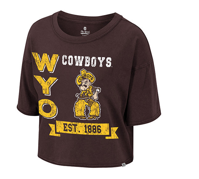 A dark brown cropped t-shirt featuring a distressed yellow and white graphic of the Wyoming Cowboys logo, including the text 'WYOMING COWBOYS', 'WY O', 'EST. 1886', and the cowboy mascot.