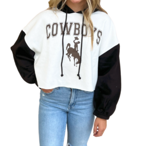 woman wearing black and white hooded sweatshirt, black sleeves, hood and drawstrings, white body, design is word Cowboys in brown with brown bucking horse below