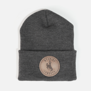 Dark grey beanie, design is brown leather patch with word Wyoming above bucking horse above cowboys