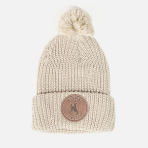 Women's cream beanie, design is brown leather patch with word Wyoming above bucking horse above cowboys