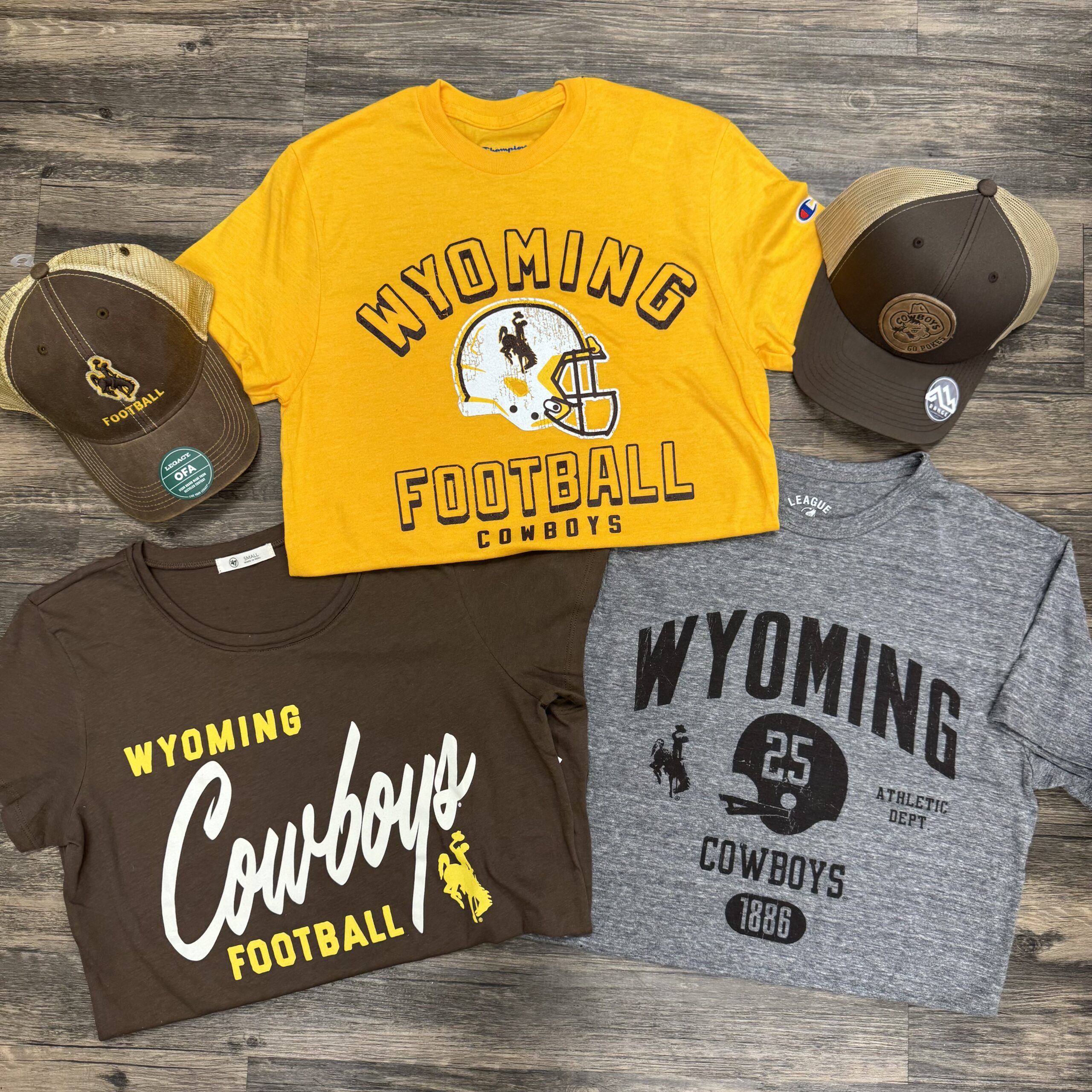 picture of a gold tee, brown tee, and a grey tee laying on the ground that say wyoming football. two hats in the picture too