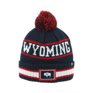 navy beanie with red and navy pom on top. has white wyoming lettering in between two red lines. has state flag patch with white and red stripes on sides on bottom of hat